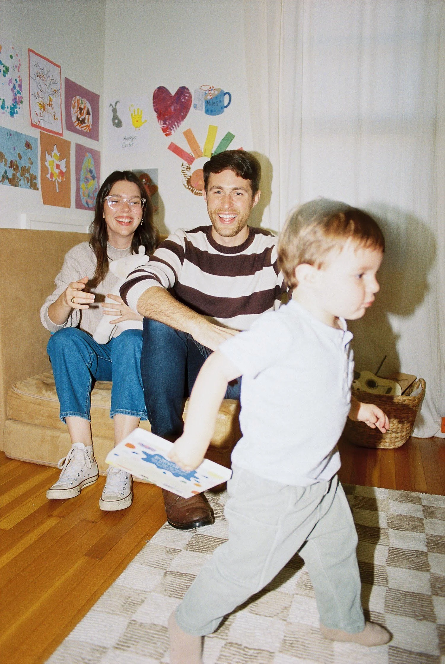A toddler hurrying past the camera with a book while two smiling grown-ups sit behind them in a room filled with children’s artwork.