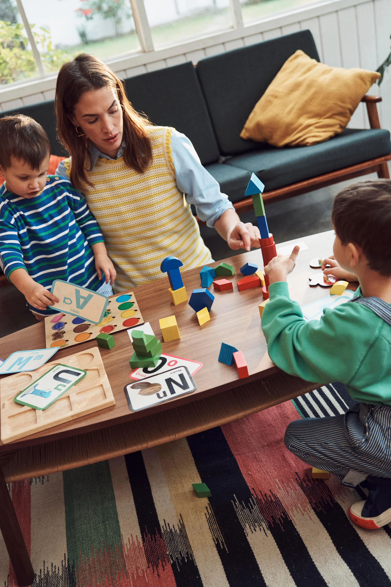 A grown-up and two young children playing with colorful blocks and cards around a table.